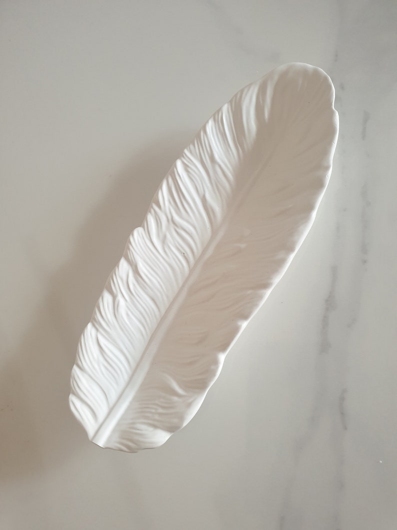 May include: White ceramic feather-shaped dish. The dish is designed to look like a real feather with detailed ridges and a smooth, glossy finish.