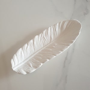 May include: White ceramic feather-shaped dish. The dish is designed to look like a real feather with detailed ridges and a central vein.