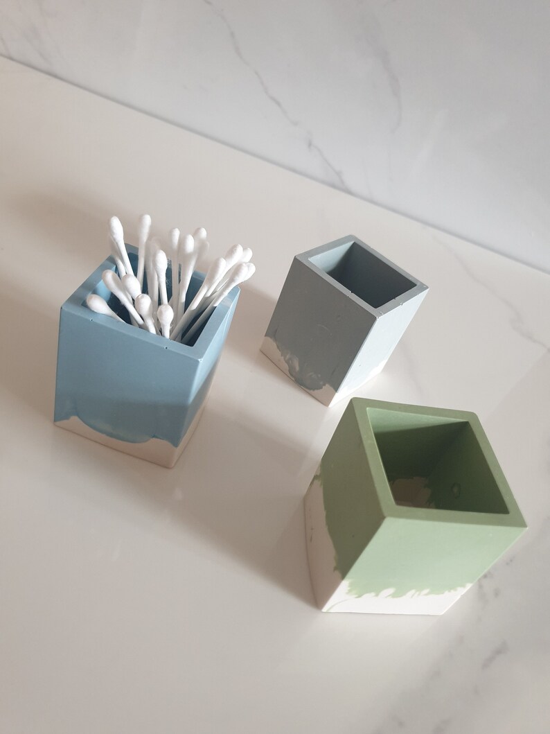 May include: Three small square concrete containers with a white base. One container is light blue, one is gray, and one is green. The light blue container has cotton swabs inside.