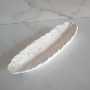 May include: White ceramic feather-shaped tray. The tray is designed to look like a real feather with detailed ridges and a smooth, glossy finish.