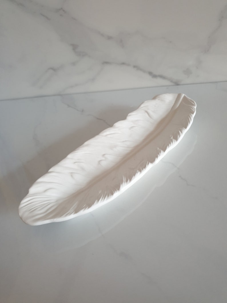 May include: White ceramic feather-shaped tray. The tray is designed to look like a real feather with detailed ridges and a smooth, glossy finish.