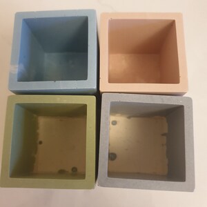 May include: Four square-shaped planters in different colors: light blue, light pink, light green, and gray. The planters are made of concrete and have a smooth finish.