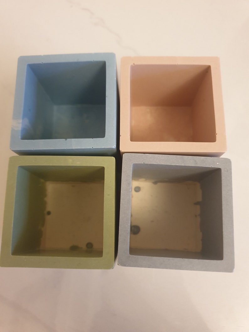 May include: Four square-shaped planters in different colors: light blue, light pink, light green, and gray. The planters are made of concrete and have a smooth finish.