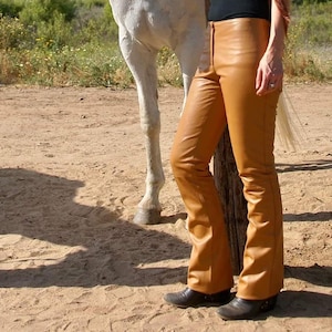 May include: A woman wearing a black vest top and brown leather trousers stands next to a white horse. The woman is holding the horse's halter and the horse is wearing a chain halter.