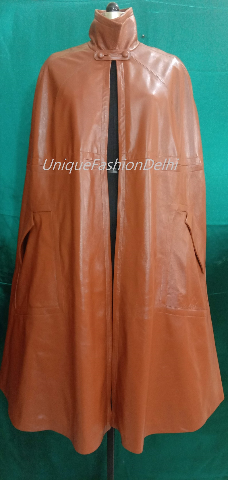 May include: A brown leather cape with a high collar and a button closure. The cape has two pockets on the sides and a long, flowing design. The text "UniqueFashionDelhi" is visible on the front of the cape.