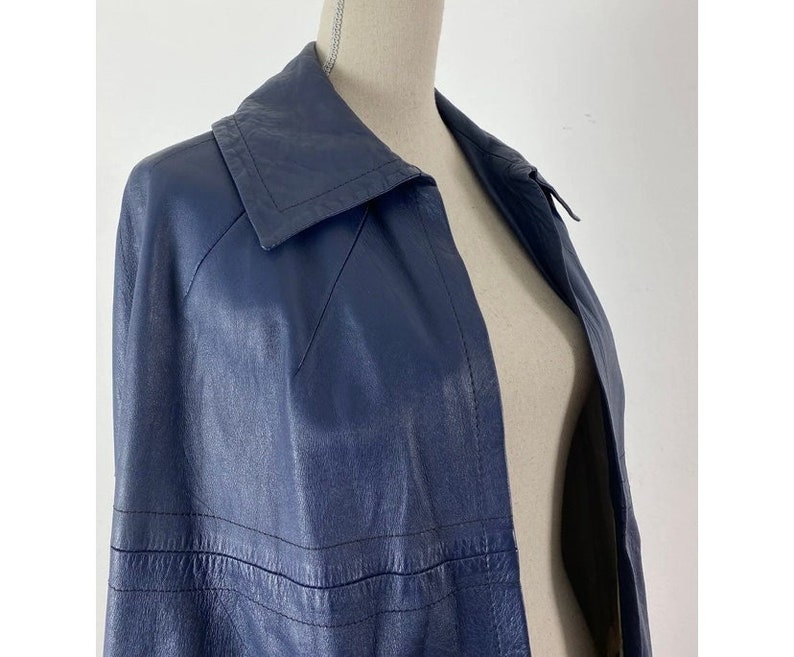 May include: A navy blue leather jacket with a long, fitted silhouette and a large collar. The jacket has a single-breasted closure and a smooth, polished finish.