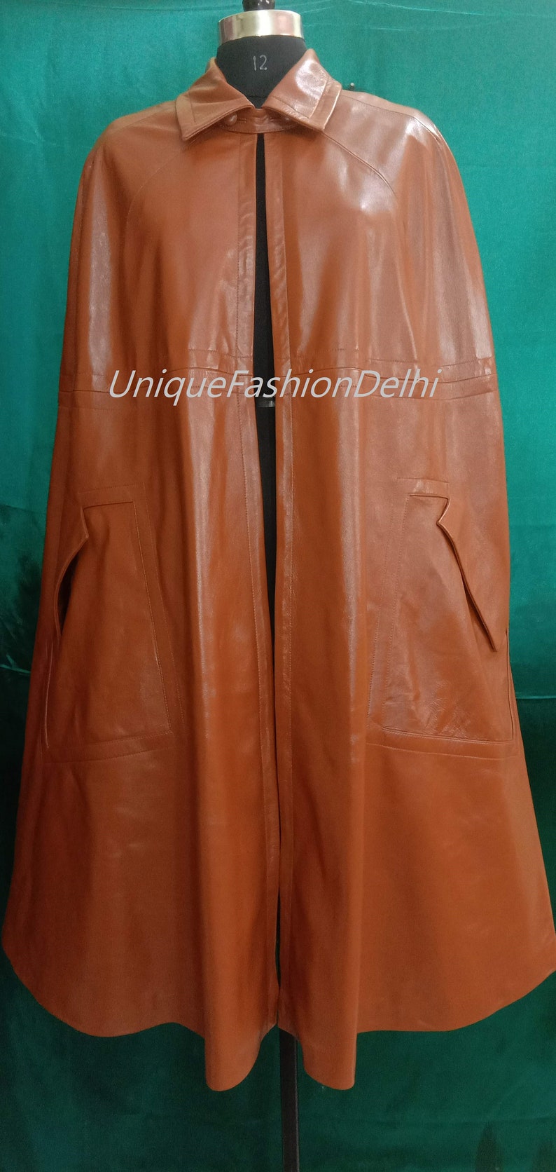May include: A brown leather cape with a collar and a long, flowing design. The cape has a slit down the back and two pockets on the sides. The text "UniqueFashionDelhi" is printed on the front of the cape.