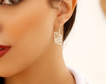 May include: A silver drop earring featuring a cutout design of intricate Persian calligraphy. The earring hangs from a simple silver hook and displays a polished metallic finish, creating a delicate and detailed appearance on the earlobe.