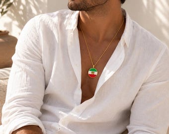 May include: A gold-colored necklace with a round pendant featuring the colors of the Mexican flag: green, white, and red. The pendant is suspended from a delicate gold chain. The person is wearing a white button-down shirt.