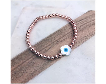 May include: A rose gold beaded bracelet with a white flower charm featuring a blue evil eye.