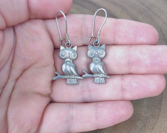 May include: A pair of silver owl earrings with a detailed design. The owls are perched on branches and have large, round eyes.