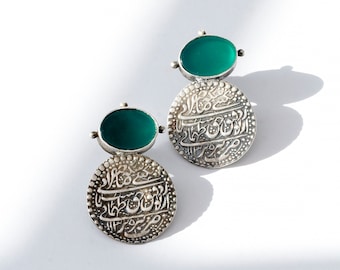 May include: A pair of silver earrings with a coin-like design. Each earring features a round, green gemstone at the top and a larger, engraved silver disc below. The discs have intricate calligraphy. The earrings are set against a white background.