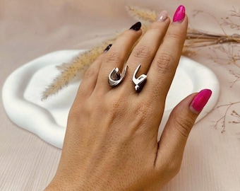 May include: Two silver rings, each featuring a unique Arabic calligraphy design, are displayed on a hand with polished nails. The rings are the focal point, showcasing a minimalist and elegant aesthetic. The background is a soft, neutral tone.