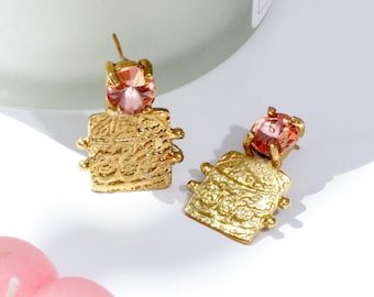 May include: A pair of gold earrings with a textured, ornate design and a pink gemstone accent. The earrings are dangling and have a square shape.