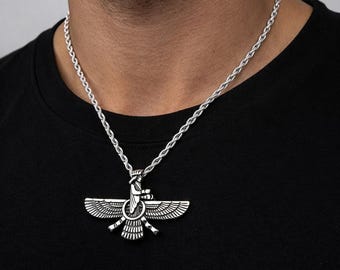 May include: Silver-toned pendant necklace featuring a detailed Faravahar symbol. The pendant is suspended from a rope chain. The necklace is worn against a black shirt.