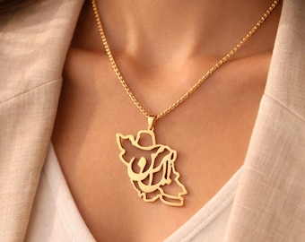 May include: Gold-colored pendant necklace featuring a stylized design. The pendant hangs from a delicate chain and is worn against a neutral-toned top and blazer. The necklace is a piece of jewelry.