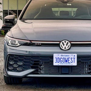 Puede incluir: Un coche Volkswagen GTI gris con una matrícula que dice "3DGOWEST" está estacionado frente a un concesionario. Otros coches están estacionados en el fondo. El cielo es una mezcla de naranja y amarillo.