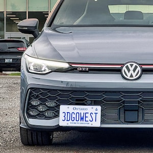 Puede incluir: Un Volkswagen Golf GTI gris visto desde el frente, con una línea roja en la parrilla, un emblema VW prominente y una placa de matrícula blanca de Ontario con el texto 3DGOWEST montada en el parachoques inferior de rejilla negra.