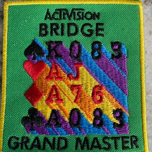 May include: A square, embroidered patch with the text "Activision Bridge Grand Master." The patch features card suits, card values, and a colorful, striped design in shades of green, yellow, purple, red, and blue.