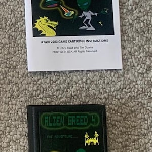 Puede incluir: Un cartucho de juego Atari 2600 y un manual de instrucciones para "Alien Greed 4". El cartucho y el manual presentan un diseño verde y negro con gráficos de extraterrestres y castillos. El texto "Alien Greed 4" se muestra de forma destacada.