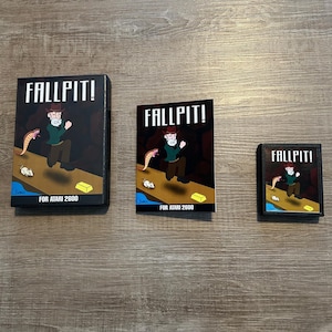 Könnte beinhalten: Drei verschiedene Versionen einer Vintage-Atari-2600-Videospielkassette. Das Spiel heißt "Fallpit!" und zeigt einen Cartoon-Mann, der in eine Grube fällt. Die Kassette ist schwarz mit einem bunten Etikett. Das Etikett zeigt einen Cartoon-Mann, der mit einer Schlange und einem Goldbarren in eine Grube fällt.