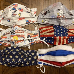 Könnte beinhalten: Sechs verschiedene Stoffmasken mit patriotischen Motiven. Die Masken sind in den Farben Rot, Weiß und Blau gehalten und zeigen Sterne, Streifen und Bilder von Snoopy.