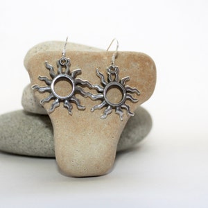 May include: A pair of silver sun earrings with a circular design. The earrings are hanging from a stone surface.