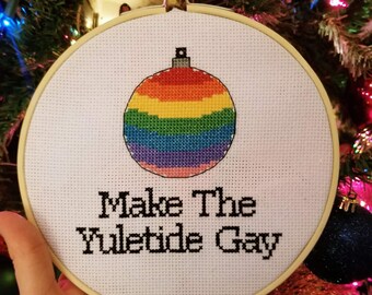 May include: A cross-stitch embroidery hoop with a rainbow bauble design. The text "Make The Yuletide Gay" is stitched below the bauble in black thread. The hoop is held in front of a Christmas tree with festive decorations.
