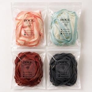May include: Four bath sponges in individual clear plastic pouches, each labeled BOCCE. Bath Sponge. The sponges come in coral, light blue, dark red, and black, featuring a textured mesh material. Each package includes icons for luxe feel, travel, and hygiene.
