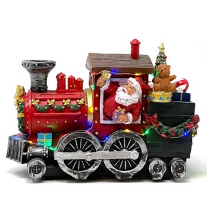 May include: A red and black Christmas train with Santa Claus riding in the caboose. The train has lights and is decorated with greenery and bows.
