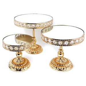 May include: Three gold-colored decorative stands with mirrored tops. The stands have ornate bases and are embellished with clear, sparkling crystals around the edges of the mirrored surfaces. The stands are of varying heights, creating a tiered display.