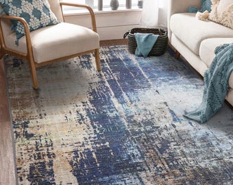 May include: A rectangular area rug with an abstract design in shades of blue, beige, and brown. The rug has a distressed, painterly appearance, with a mix of light and dark tones creating a textured effect. The rug is placed in a living room setting.