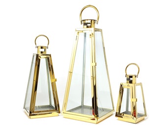 May include: Three gold metal lanterns of different sizes with glass panels. The lanterns are pyramid shaped with a handle on top.
