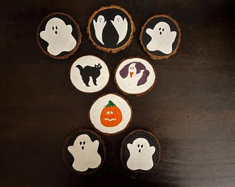 May include: Eight wooden coasters with Halloween-themed designs. The coasters feature painted images of ghosts, a black cat, a pumpkin, and a dog. The coasters are all round and have a natural wood finish.