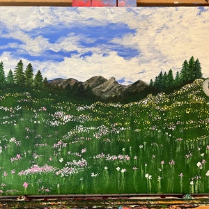 May include: Acrylic painting depicting a colourful landscape with a meadow of wildflowers in pink, white, and yellow. The scene includes a mountain range, evergreen trees, and a winding path under a blue sky with white clouds. The artwork is on a canvas.