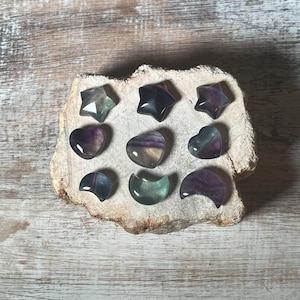 May include: Nine fluorite crystal carvings in the shapes of stars, hearts, and crescent moons. The crystals are a mix of purple, green, and black.