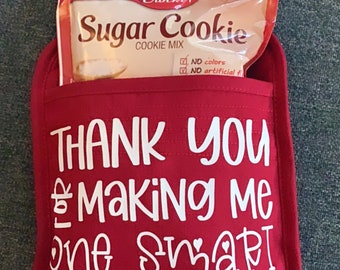 Op de afbeelding: Een rode ovenwant met witte tekst die zegt "Bedankt dat je me een slimme koekje hebt gemaakt". Een zak Betty Crocker Sugar Cookie mix zit in de zak van de ovenwant.