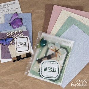 May include: A clear plastic bag filled with assorted paper ephemera, including patterned paper, stickers, and a butterfly. The bag is sitting on a brown surface with a stack of different coloured paper sheets behind it.