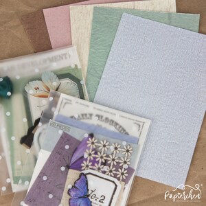 May include: A set of decorative papers with a variety of textures and colours, including brown, pink, cream, green, and blue. The papers are arranged in a stack, with the top paper being a light blue textured paper. The papers are ideal for scrapbooking, card making, and other craft projects.