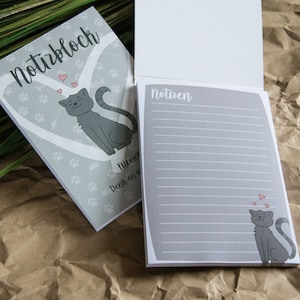 Könnte beinhalten: Grauer Notizblock mit einer Katzenillustration und dem Text "Notizblock" auf dem vorderen Umschlag. Der hintere Umschlag hat den Text "Notizen" und liniertes Papier zum Schreiben von Notizen.