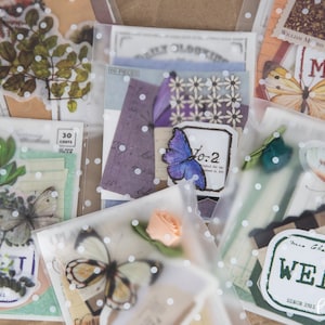 May include: Assortment of clear plastic bags with white polka dots, each containing various decorative paper ephemera, including butterflies, flowers, and leaves. The bags are labelled with the days of the week, "Mon", "Wed", and "To-2".