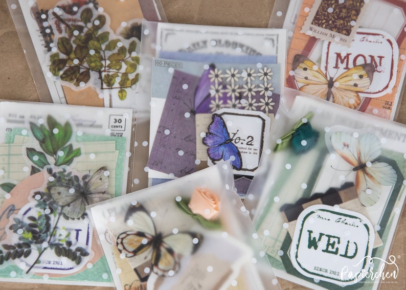 May include: Assortment of clear plastic bags with white polka dots, each containing various decorative paper ephemera, including butterflies, flowers, and leaves. The bags are labelled with the days of the week, "Mon", "Wed", and "To-2".