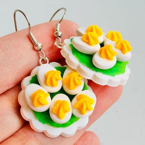 May include: Handmade novelty earrings featuring miniature deviled eggs. Each earring showcases a white plate with a green base, topped with egg halves and yellow filling. The earrings have silver-colored fish hook ear wires.