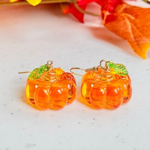 May include: A pair of orange glass pumpkin earrings with green leaf accents. The earrings are on a white surface.