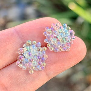 May include: Two iridescent stud earrings. Each earring is composed of numerous small, clear, bubble-like shapes. The bubbles reflect a spectrum of colors, including pink, purple, and blue. The earrings are set against a blurred green background.