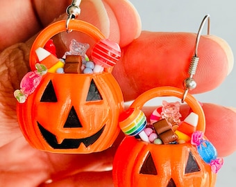 May include: A pair of orange jack-o'-lantern bucket earrings featuring black facial features and filled with miniature colourful sweets, including candy corn, lollipops, and small chocolate bars, attached to silver-toned metal earring hooks.