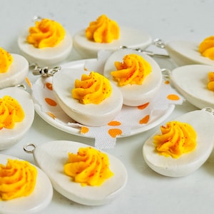 May include: A collection of white ceramic egg-shaped earrings with yellow and red yolk-like fillings. The earrings are displayed on a white surface with a white and yellow patterned fabric.