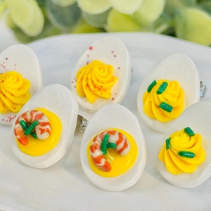 May include: Six deviled eggs with different toppings, including shrimp, yellow and green swirls, and a red and white swirl. The eggs are white with a yellow yolk and are arranged on a white plate.