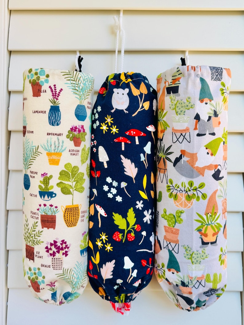 Op de afbeelding: Drie herbruikbare boodschappentassen hangen aan een witte deur. De tassen zijn gemaakt van stof en hebben verschillende patronen. De eerste tas heeft een witte achtergrond met een verscheidenheid aan planten en bloemen in potten. De tweede tas heeft een donkerblauwe achtergrond met witte bloemen, rode paddenstoelen en aardbeien. De derde tas heeft een witte achtergrond met een verscheidenheid aan planten en dwergen.
