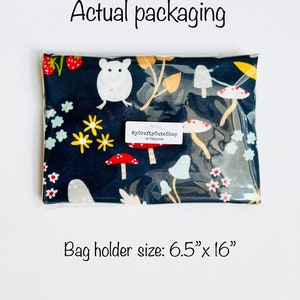 Op de afbeelding: Een doorzichtige plastic zak met een zwarte stoffen tas erin. De tas heeft een witte muis, rode en witte paddenstoelen en gele en groene bladeren op een donkerblauwe achtergrond. De zak is gelabeld met "MyCraftyCuteShop at Etsy.com". De maat van de tasdrager is 16,5 cm x 40,6 cm.